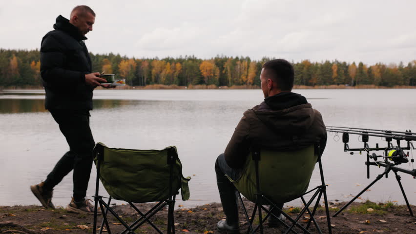 Two fishermen relaxing by lake in autumn, sharing food and conversation near fishing rods, peaceful outdoor leisure with friendship and camping atmosphere in calm nature setting