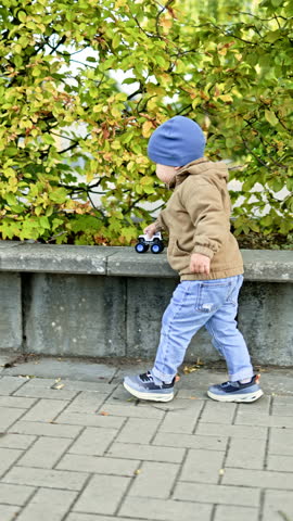 Little 2 year old boy playing with a toy car outside. Child playing outdoors. Toddler in jacket and hat playing outside.