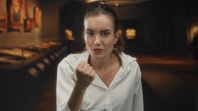 Young hispanic woman clenches fists and frowns while standing in dimly lit art gallery building hallway lined with framed paintings; anger. - Powered by Shutterstock - Get 15% off with code: PIKWIZARD15