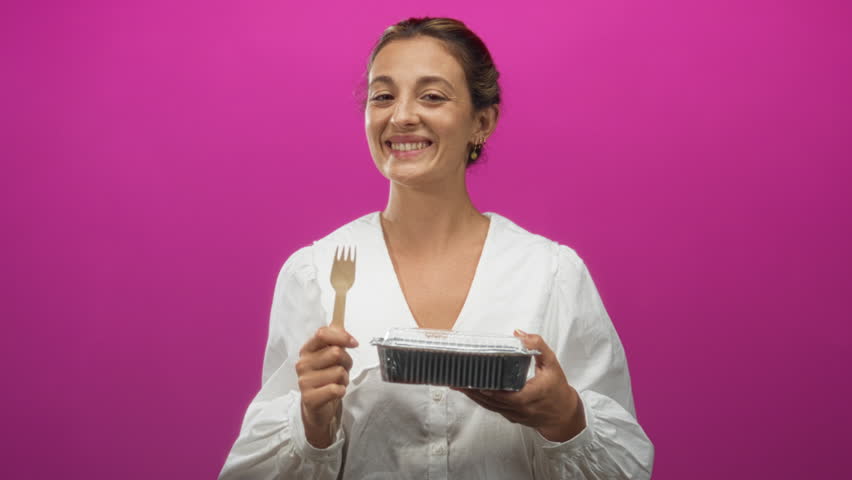 Woman smiling and holding a wooden fork and aluminum takeaway tray while wearing white blouse in pink studio; happy appetite.