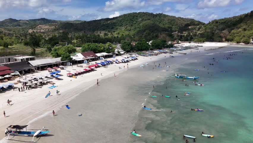 Gorgeous aerial view of Selong Belanak beach, Lombok with colorful umbrellas, swimmers, surfers and traditional boats along turquoise water and white sandy coastline