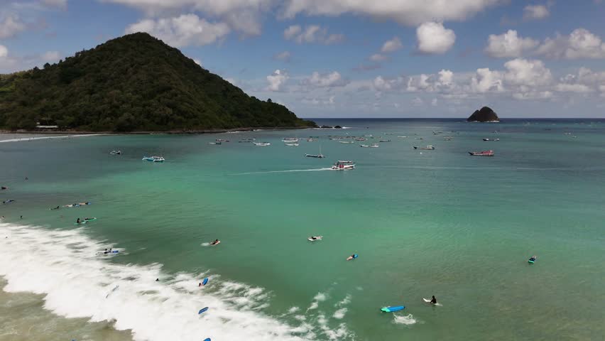 Aerial view of surfers enjoying the waves in a beautiful tropical bay with turquoise water, lush green hills, and traditional fishing boats dotting the coastline under a partly cloudy sky