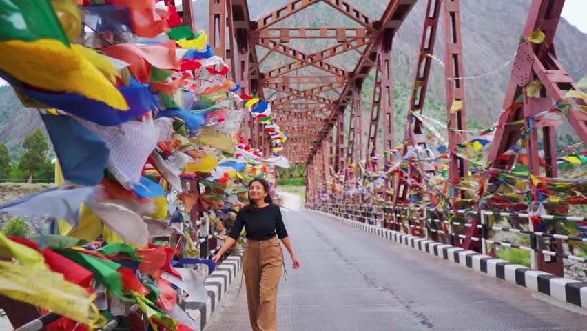  Indian girl walk near colorful prayer flags hanging on metal bridge in mountain valley near Mini Manali in Lahaul, Himachal Pradesh, India. Female tourist exploring remote village of Himalayas. 