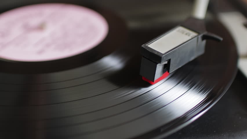 Close-up of a vinyl record playing on a turntable. The stylus on the tonearm follows the grooves of the spinning black disc with music