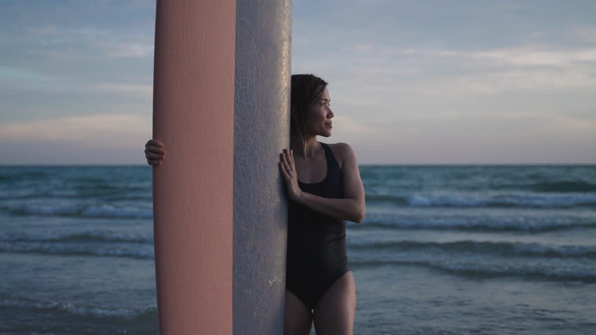 Portrait of a happy Asian woman surfer in a bikini standing smiling and holding a surfboard while looking at the camera outdoors on summer vacation during sunset on the beach after surfing. 