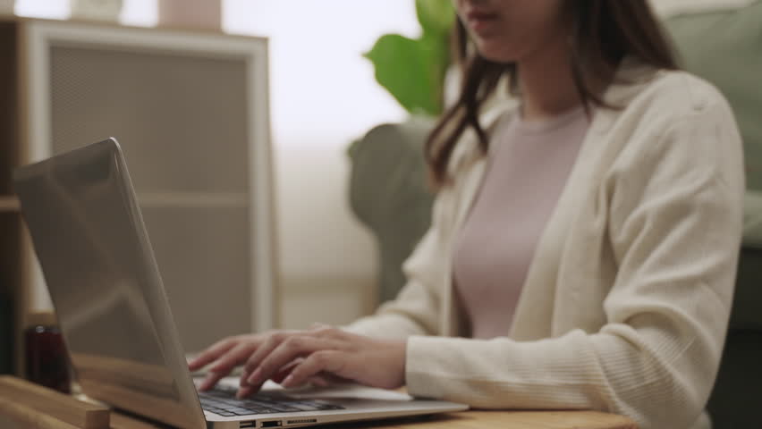 Selection focuses on hands. Close up of a young Asian woman sitting and typing a document report on the keyboard of a laptop computer about working and online learning in the living room at her home. 