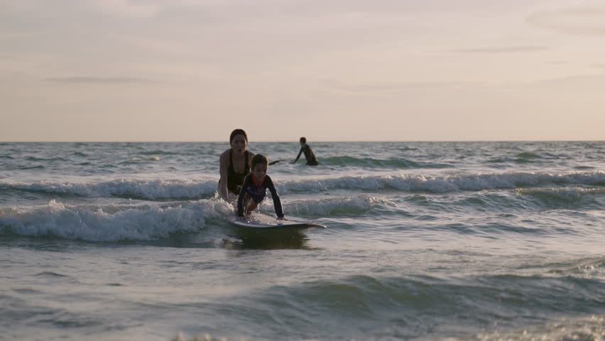 Happy little boy learning to play with a surfboard from his mother, having fun and passion in the sea during summer vacation at the beach. An Asian mom teaches and trains her cute son on how to surf. 