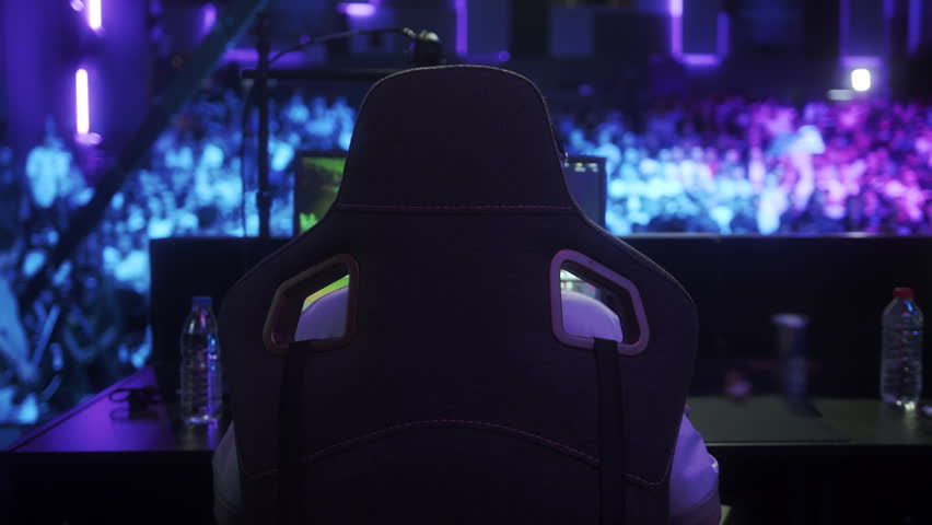 Professional esports player sitting in a gaming chair on stage facing the audience, focused on gameplay under colorful tournament lights.