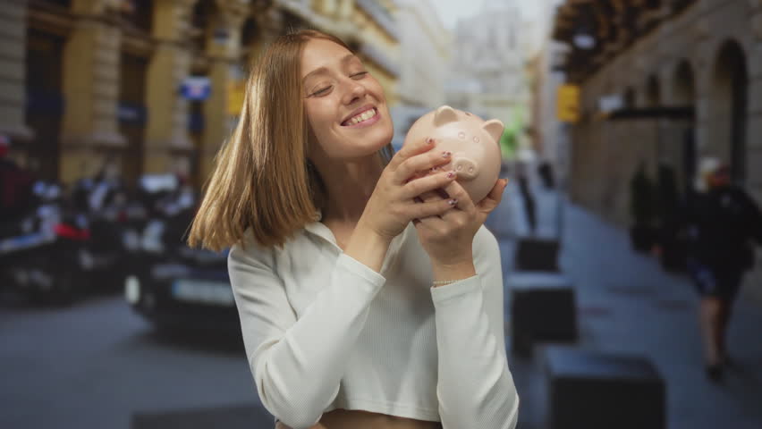 Young blonde woman smiling and kissing a piggy bank on urban street with taxi in background, showcasing city life and savings concept.