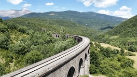 Curving train tracks arching over stone viaduct bridge spanning verdant mountain valley beneath bright summer sky, showcasing scenic railway infrastructure - Powered by Shutterstock - Get 15% off with code: PIKWIZARD15