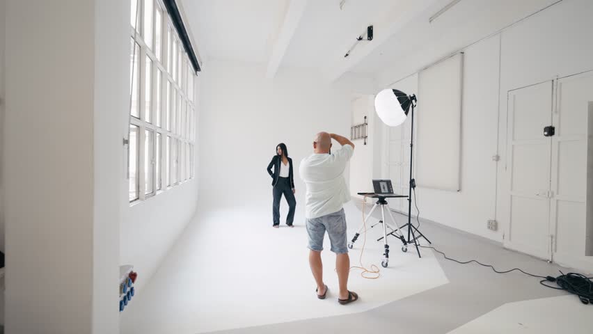Professional photographer capturing sleek business portrait of confident businesswoman posing near white studio walls with professional lighting and camera equipment