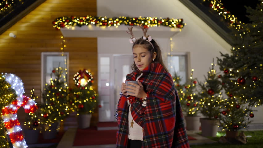 A young girl wrapped in a blanket sips from a mug in front of a house decorated for Christmas