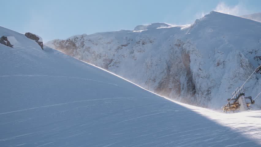Snow blowing in the wind over a sunlit mountain peak with a ski lift in view. A breathtaking winter scene showing the power and beauty of nature in cold conditions.
