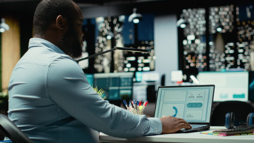 Black advisor researching market data and building strategy for business targets, using spreadsheets on laptop for proofreading. Late night overtime for a multinational company. Camera B.