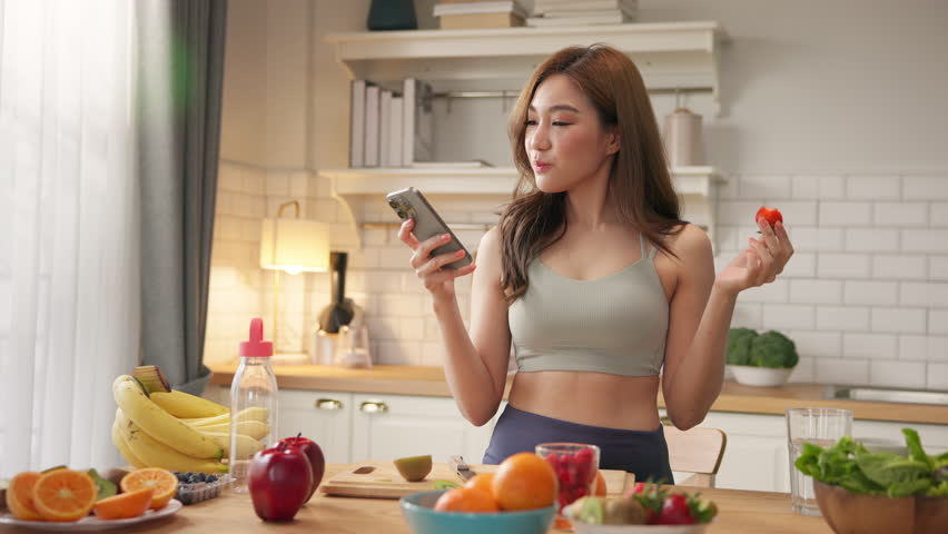 Young Asian woman in sportswear enjoys organic healthy fruits in kitchen while checking health data on smartphone, using AI-powered nutrition or wellness app to support clean diet balanced lifestyle.