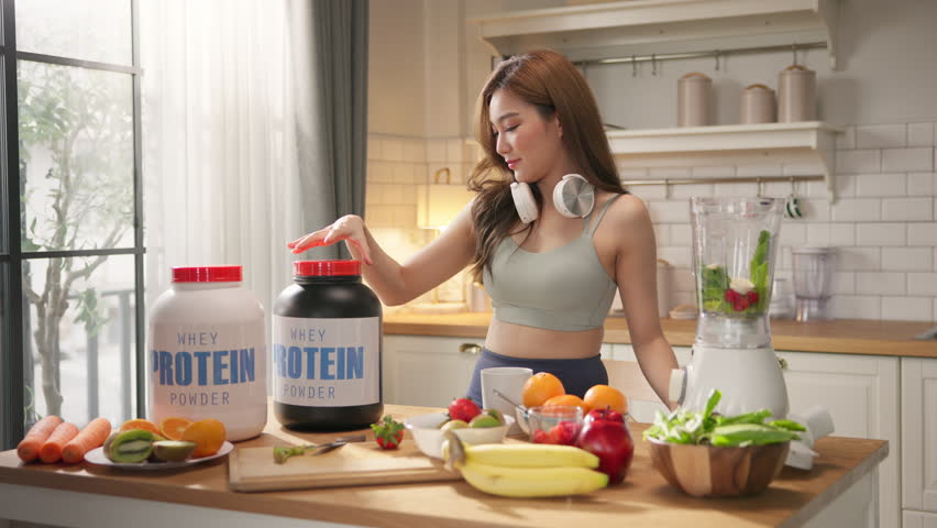 Young Asian woman in sportswear making healthy protein shake smoothie in home kitchen, using organic fresh fruits, vegetables, and whey powder. Wellness, fitness lifestyle, balanced diet, weight loss.