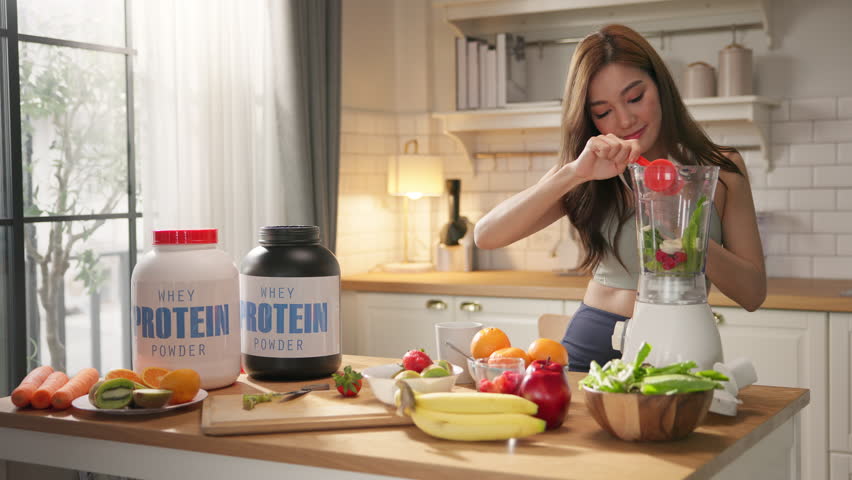 Young Asian woman in sportswear making healthy protein shake smoothie in home kitchen, using organic fresh fruits, vegetables, and whey powder. Wellness, fitness lifestyle, balanced diet, weight loss.