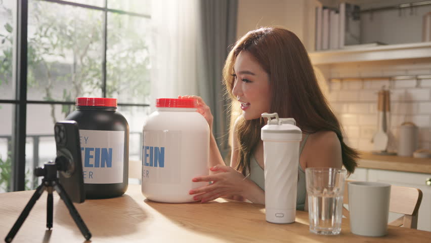 Young Asian female health influencer live streaming on social media sharing nutrition tips with whey protein powder by smartphone. Healthy lifestyle, fitness coaching, clean eating. online marketing.
