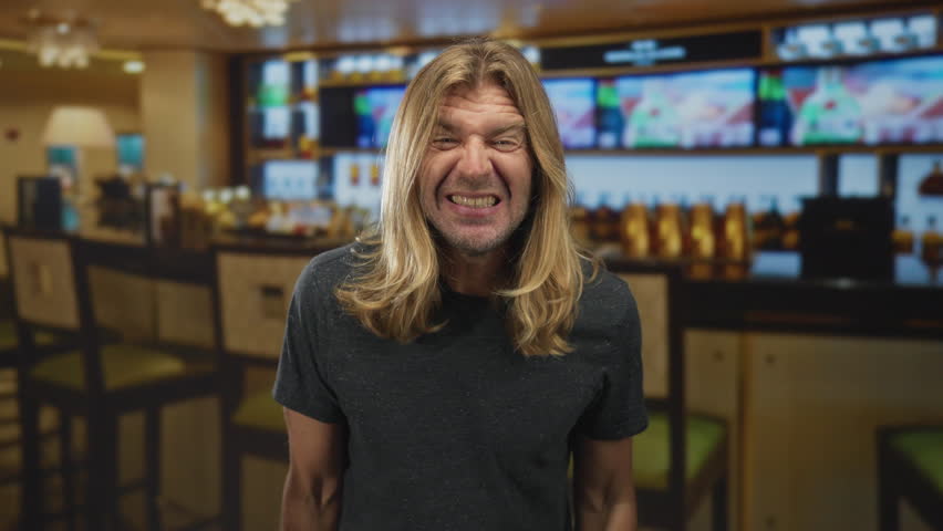 Man with long blond hair wearing casual shirt holding hand to mouth as he shouts across restaurant tables; excitement.