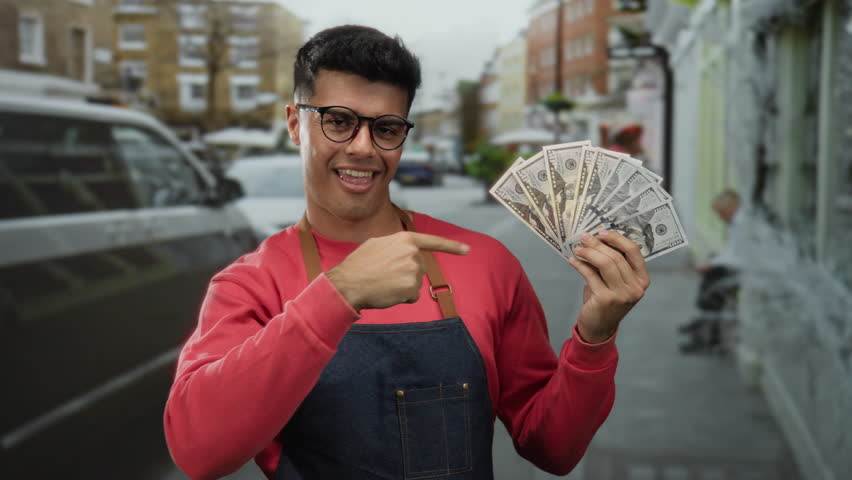 Man outdoors in city holding american banknotes, wearing glasses and apron, smiling and gesturing confidently on street.