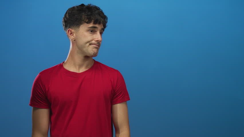 Man pointing finger while wearing a red tshirt in a blue studio, standing slightly turned with casual posture and direct gaze; doubt.