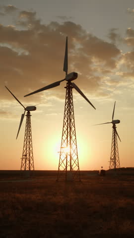 Silhouettes of wind turbines generator power plant towers against background of sunset sky with sun, vertical video