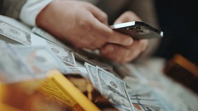 Closeup of a rich businessman's hands using a smartphone on a table covered with stacks of hundred-dollar bills and gold bars, symbolizing wealth, investment, and financial success - Powered by Shutterstock - Get 15% off with code: PIKWIZARD15