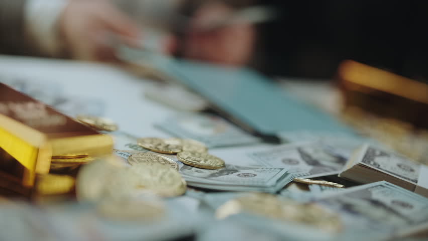 Unrecognizable person counting us dollar banknotes on a table covered with cash, physical bitcoins, and gold bars, representing wealth, investment, and cryptocurrency trading concepts