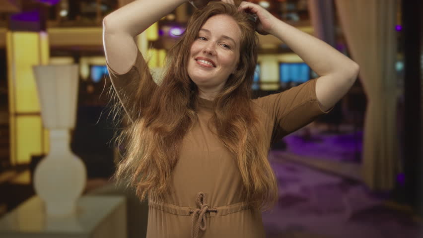 Young woman with red hair shapes heart with hands near face in hotel lobby while smiling warmly; love.