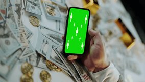 Close-up of a businessman's hands holding and using a smartphone with a chroma key green screen, displaying market data over a pile of hundred-dollar bills and gold coins - Powered by Shutterstock - Get 15% off with code: PIKWIZARD15
