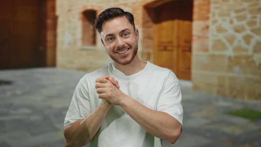 Young man with beard smiling and clasping hands outside on stone street against rustic building backdrop representing positive urban outdoor atmosphere.