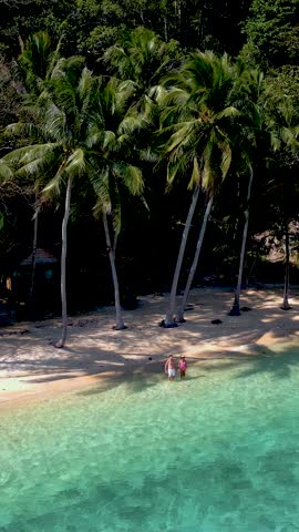Soft waves lap on the pristine sand as two travelers walk along a secluded beach in Thailand. Tall palm trees sway gently in the warm breeze, creating a peaceful paradise of Koh Wai island