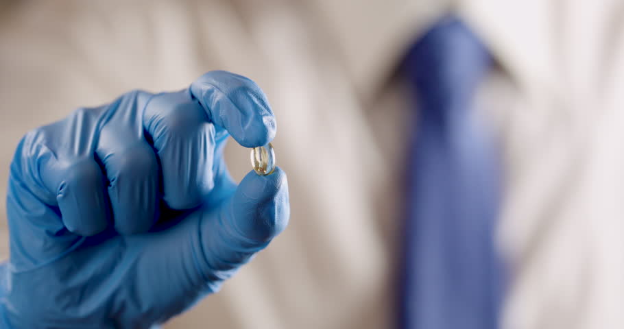  a hand wearing a blue medical exam glove, holding a golden, translucent gel capsule or supplement. The reflective surface of the pill shows a miniature, reflection of the person as the camera tracks