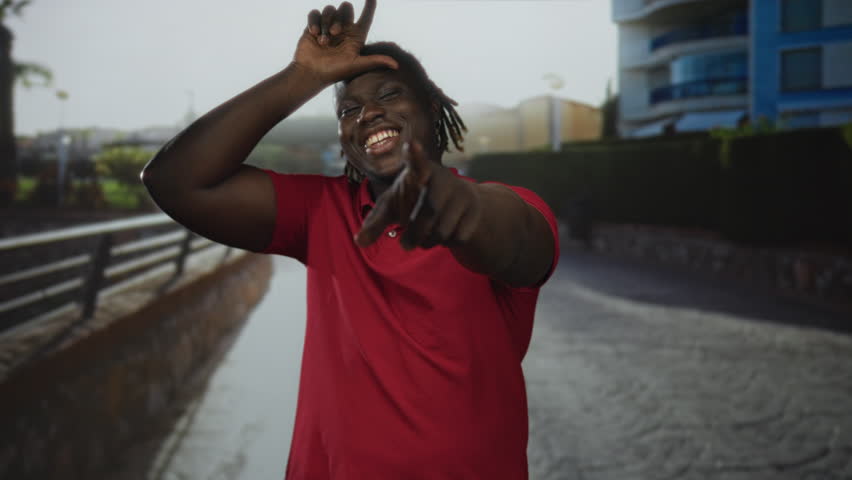Man makes l sign with hand on forehead on city street while smiling and leaning forward; playful confidence.