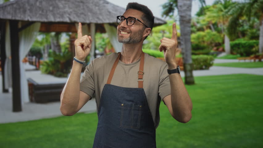 Man smiling and pointing both index fingers upward near a thatched gazebo in a forest building setting, wearing denim apron and glasses; joy welcome celebration.