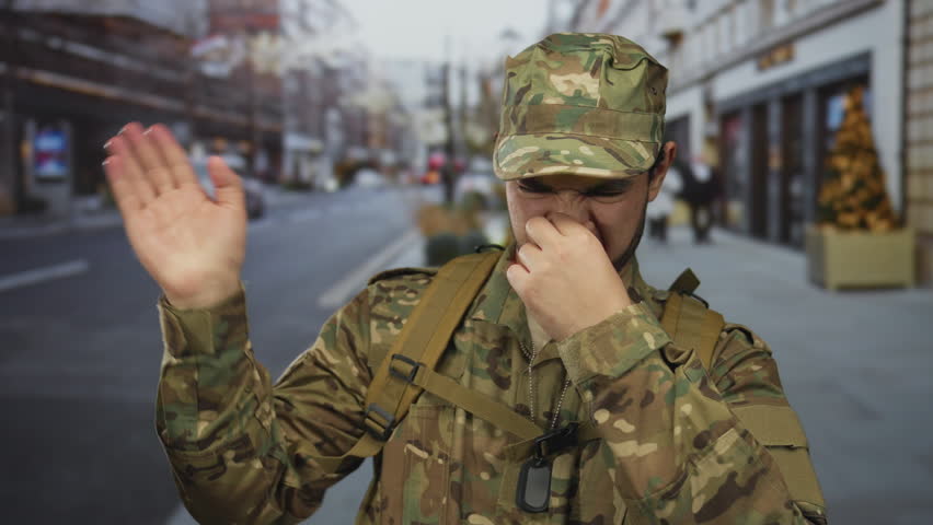 Man wearing camouflage uniform with backpack and dogtags pinches nose on busy city street; disgust.