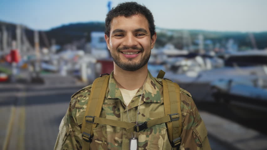 Man in camouflage uniform and backpack smiles with dog tags on sunlit port dock beside moored naval boats; pride duty camaraderie readiness.