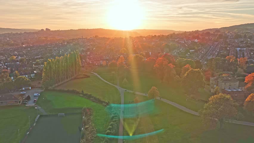 Drone pulls out above Hillsborough Park in Sheffield, South Yorkshire, England, revealing football fields, tree-lined avenues, glowing autumn foliage, and city rooftops merging into the warm sunset.