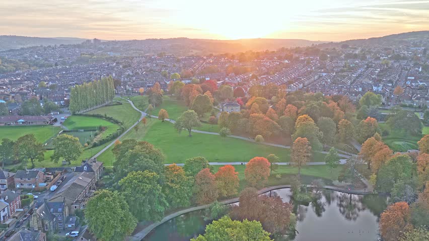Golden aerial shows Hillsborough Park in Sheffield, South Yorkshire, England, with its autumn trees, winding paths, pond, and city rooftops glowing beneath a sunset sky over this urban oasis.