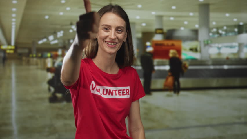 Woman volunteer giving thumbs up gesture at airport terminal wearing red volunteer shirt; community service optimism.