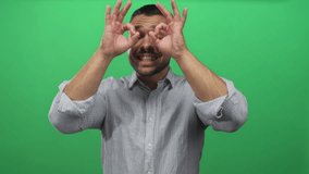 Young hispanic man in striped shirt making hand binoculars over smiling face in green studio; playful curiosity. - Powered by Shutterstock - Get 15% off with code: PIKWIZARD15