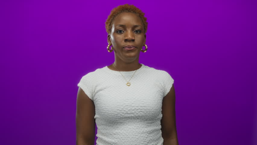 Woman wearing gold hoop earrings covering mouth with hands and wide eyes in purple studio; surprise.
