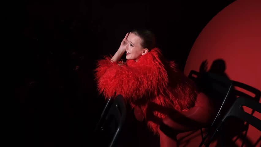 Portrait of a beautiful young woman in a red fur coat against a red background, posing for a cinema-style photo. The woman smiles beautifully, sitting on chairs in the light of a projector during a
