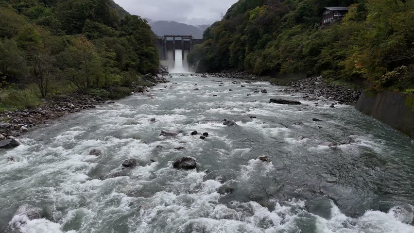 A river with a waterfall and a bridge in the background. The water is rushing and the sky is cloudy
