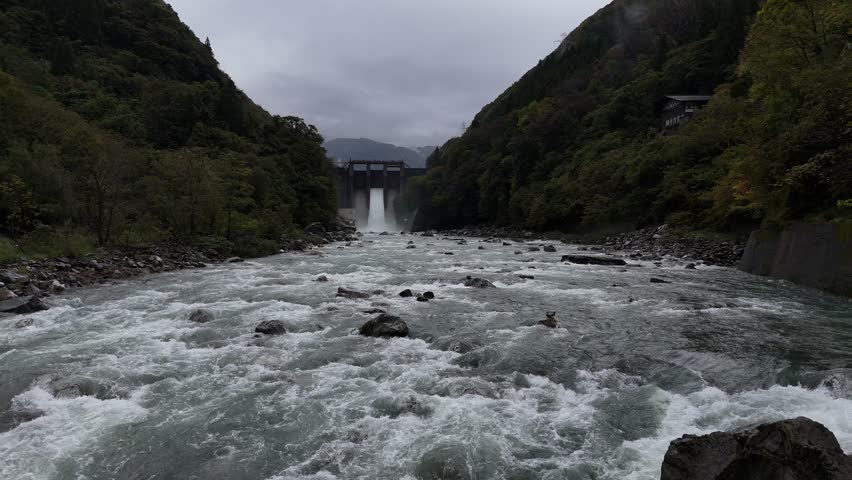 A river with a waterfall and a bridge in the background. The water is rushing and the sky is cloudy