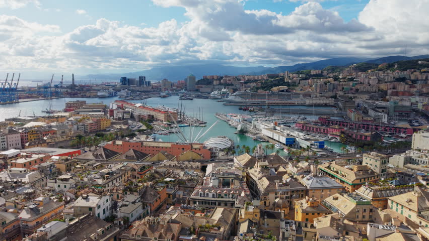 Drone flying backward over Genoa’s historic center, revealing the port, ships, and maritime buildings under a sunny sky with clouds.