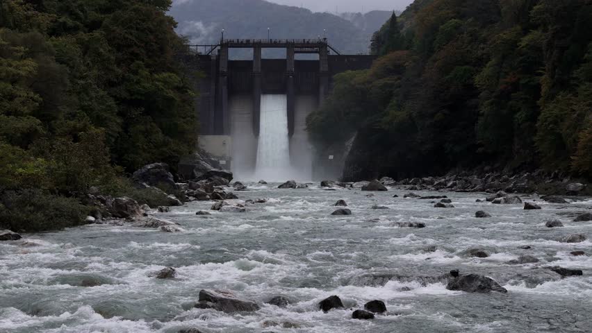 A river with a waterfall and a bridge in the background. The water is rushing and the sky is cloudy