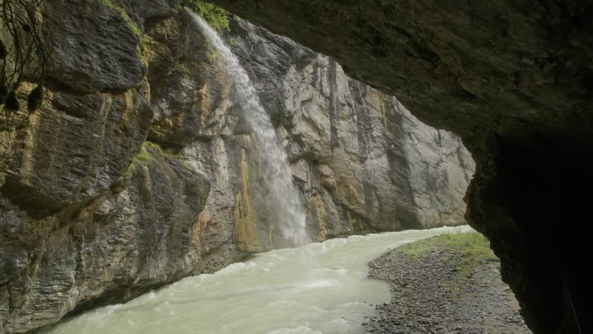 Waterfall cascading into a gorge river in the Swiss Alps, viewed from inside a cave. Dramatic mountain landscape and scenic nature scene