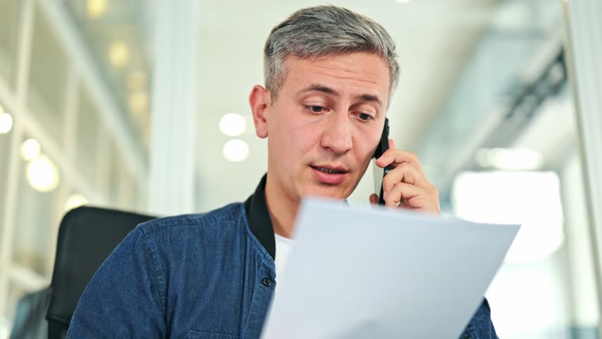 Focused businessman, dressed in a blue jacket, engages in a phone call while reviewing documents in a modern office. Concentrated and attentive, the male professional discusses important information. - Powered by Shutterstock - Get 15% off with code: PIKWIZARD15