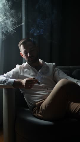 Stylish man smoking cigar indoors in low light, relaxed confident male in white shirt sitting on armchair with smoke swirling around, luxury lifestyle and masculine elegance concept